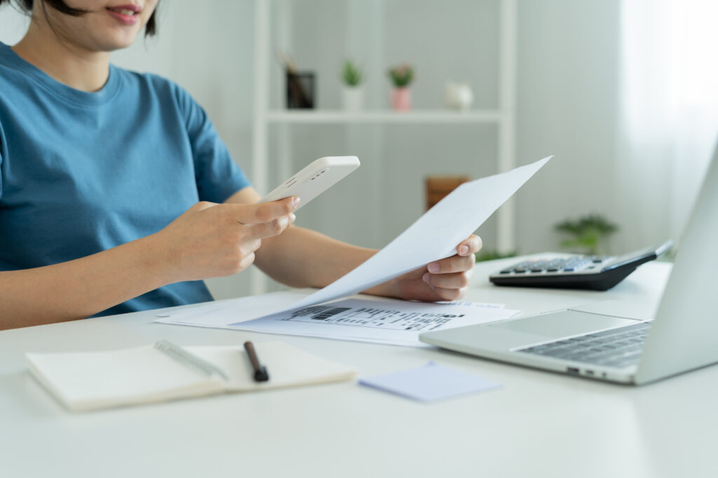 Photo of a woman sitting in front of a desk. On the desk, there is a laptop and some documents. The woman is holding one of the documents in her hand and scanning it with her smartphone.